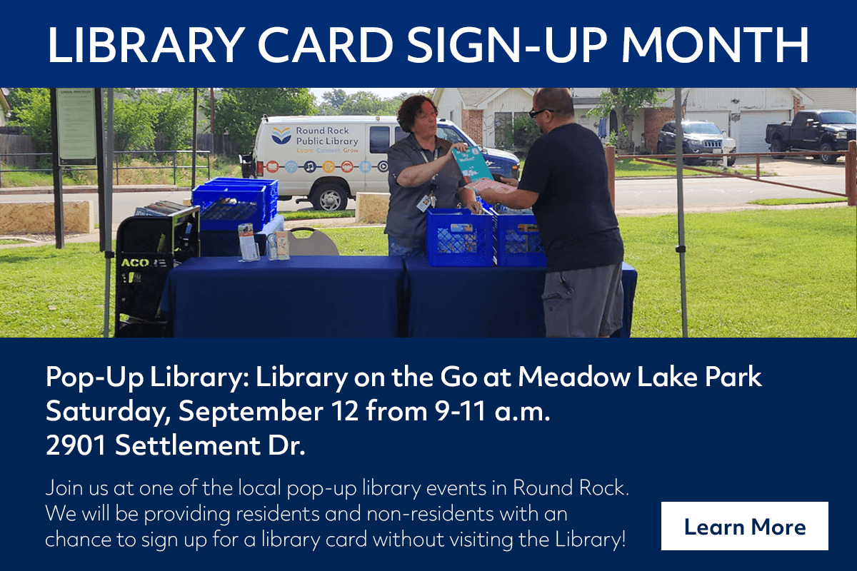 Two people stand at a table with library materials at a pop-up library event in a park, with a sign reading Library Card Sign-Up Month and event details below.