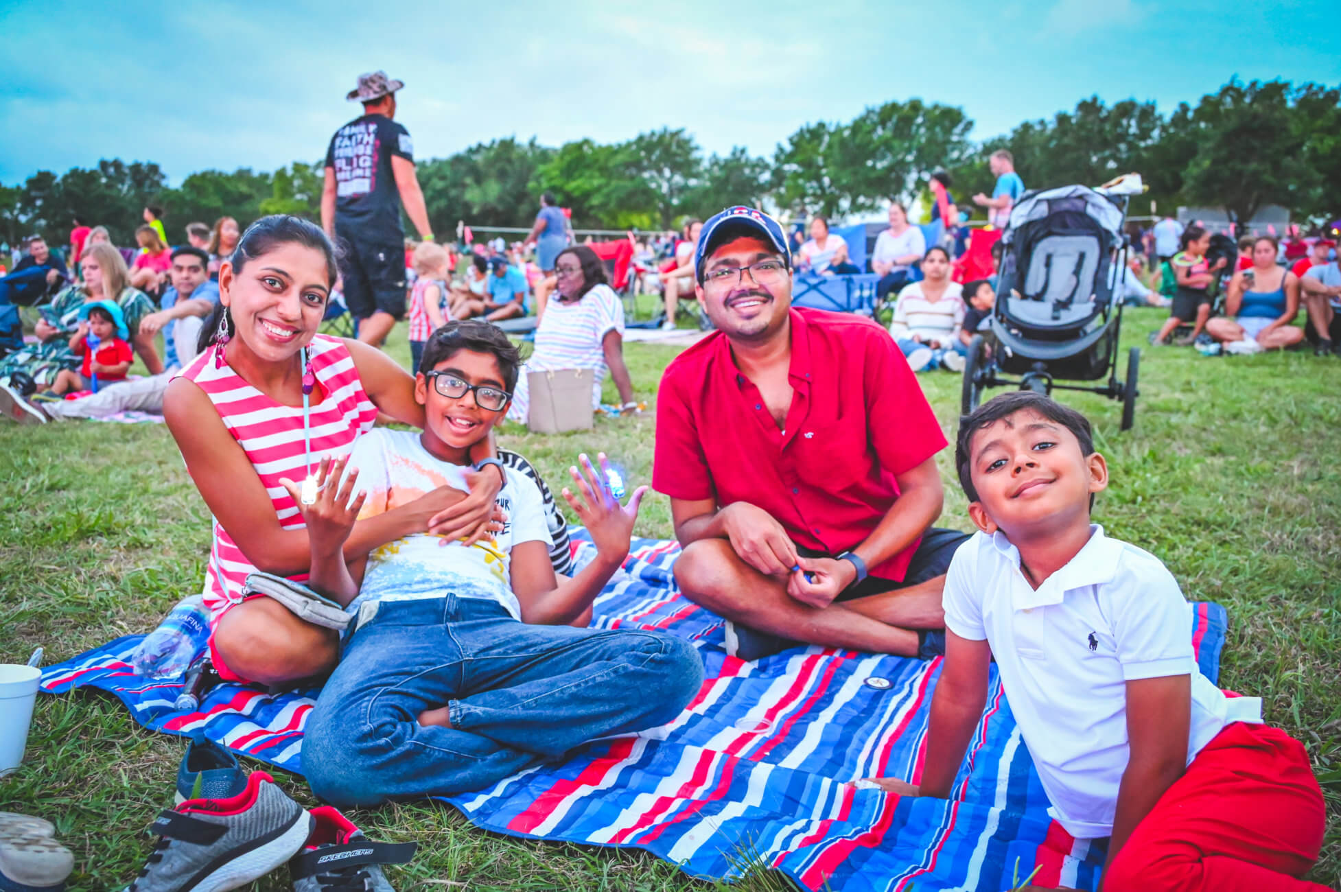 A man, woman and their two sons smile at the camera while sitting on a red, white and blue blanket at Old Settlers Park. A large crowd is seen in the background. 
