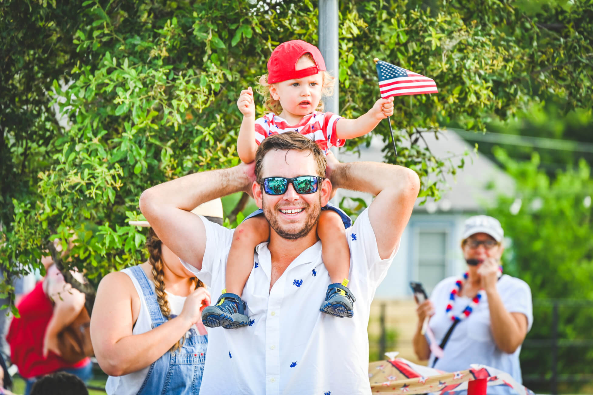 A smiling man with sunglasses and a white shirt gives his young child a piggyback ride. The child waves a small American flag. 