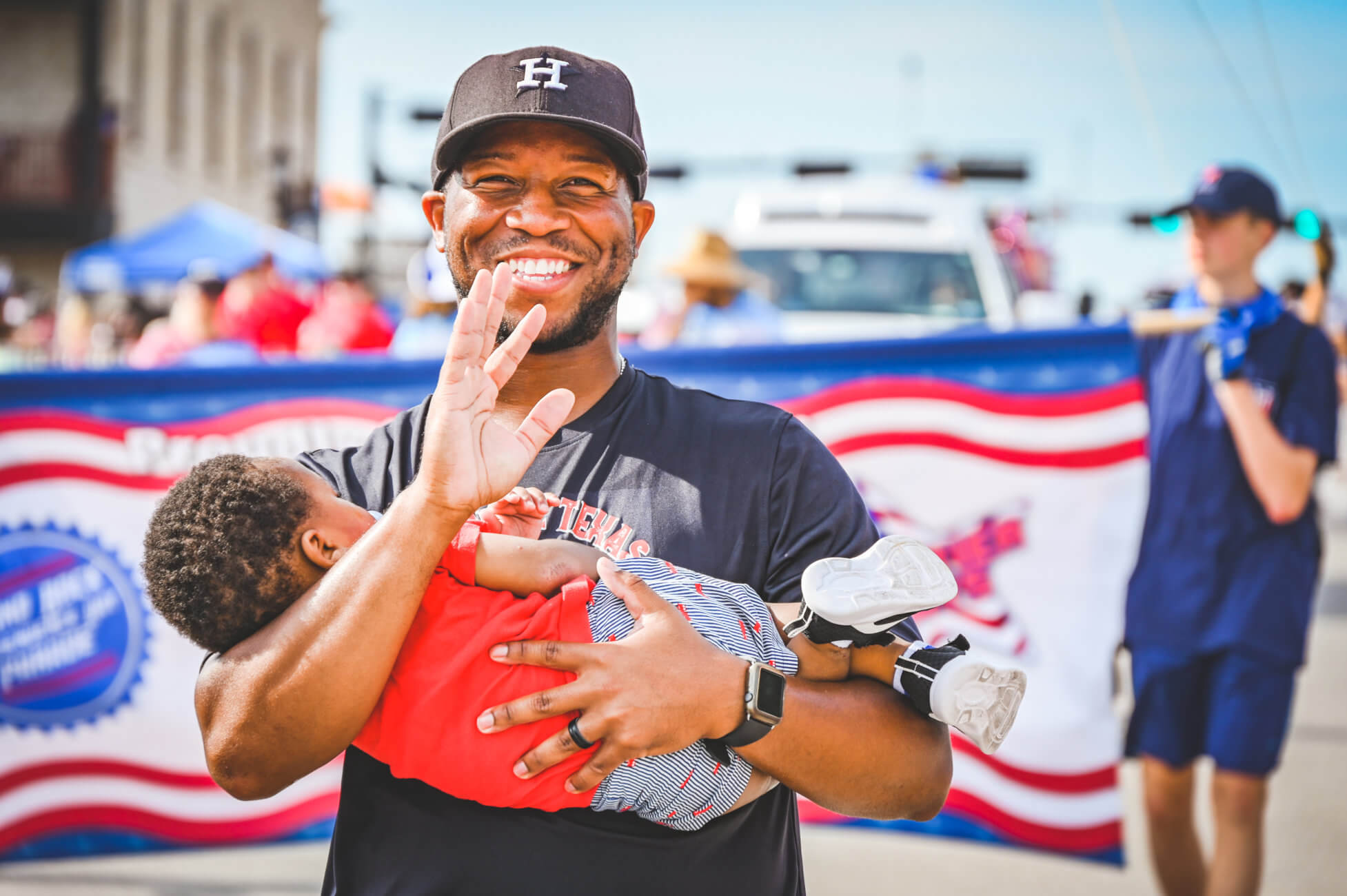 Smiling man in black baseball cap and black shirt waves to the camera while holding his young child. The parade can be seen in the background. 