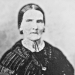 Black-and-white portrait of a woman with dark hair parted in the middle, wearing a dark dress with a lacy white collar and a ribbon tied at her neck, looking directly at the camera.