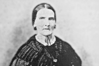 Black-and-white portrait of a woman with dark hair parted in the middle, wearing a dark dress with a lacy white collar and a ribbon tied at her neck, looking directly at the camera.