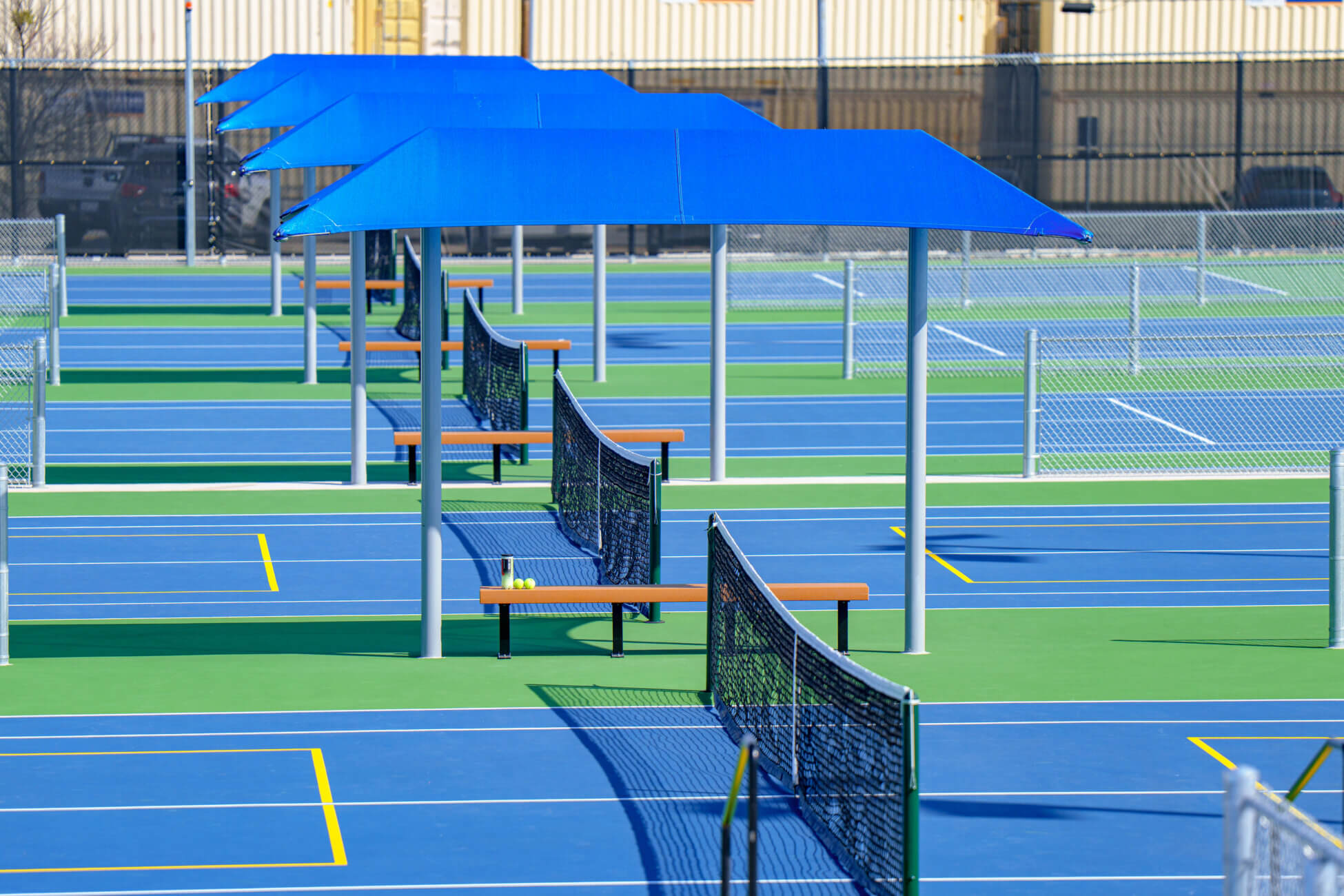 Rows of outdoor blue pickleball courts with black nets, yellow boundary lines, and blue shade structures over benches. The area is surrounded by a chain-link fence and industrial buildings are visible in the background.