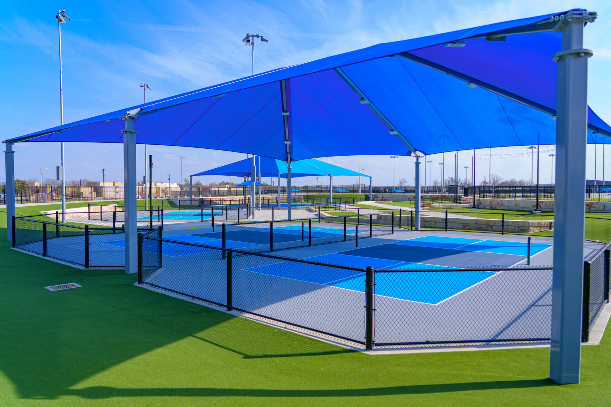 Outdoor pickleball courts with blue and turquoise surfaces are shaded by large blue canopies. The courts are surrounded by a black chain-link fence and set on green artificial turf under a sunny sky.