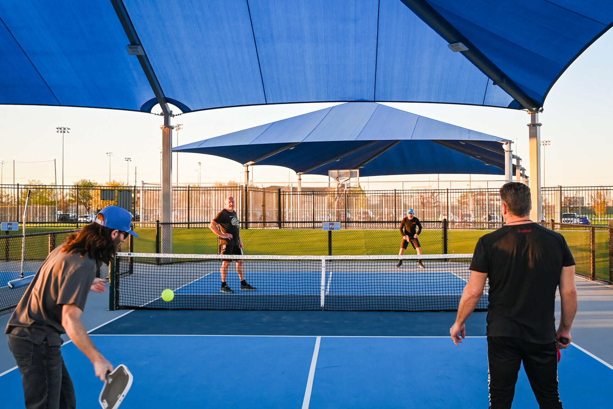 Four people play pickleball on an outdoor court under large blue shade structures. Two players are on each side of the net, with a tennis ball in mid-air. A grassy field and sports lights are visible in the background.