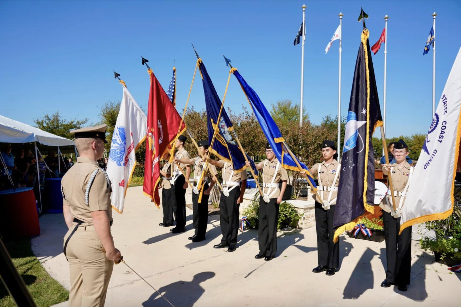 A group of uniformed military personnel stands in formation, each holding different flags, while an officer faces them. Several flagpoles and a clear blue sky are visible in the background.