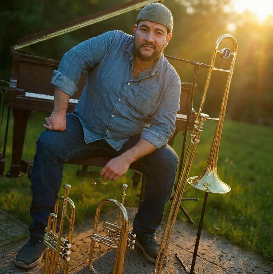 A man in a gray cap and denim shirt sits on a piano bench outdoors at sunset, with a trombone, trumpet, and flugelhorn placed in front of him on the ground.