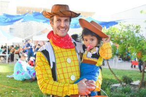 An adult and child, both dressed in matching cowboy costumes, smile at an outdoor event. The child has a pacifier and is held by the adult. There are people and tents in the background.