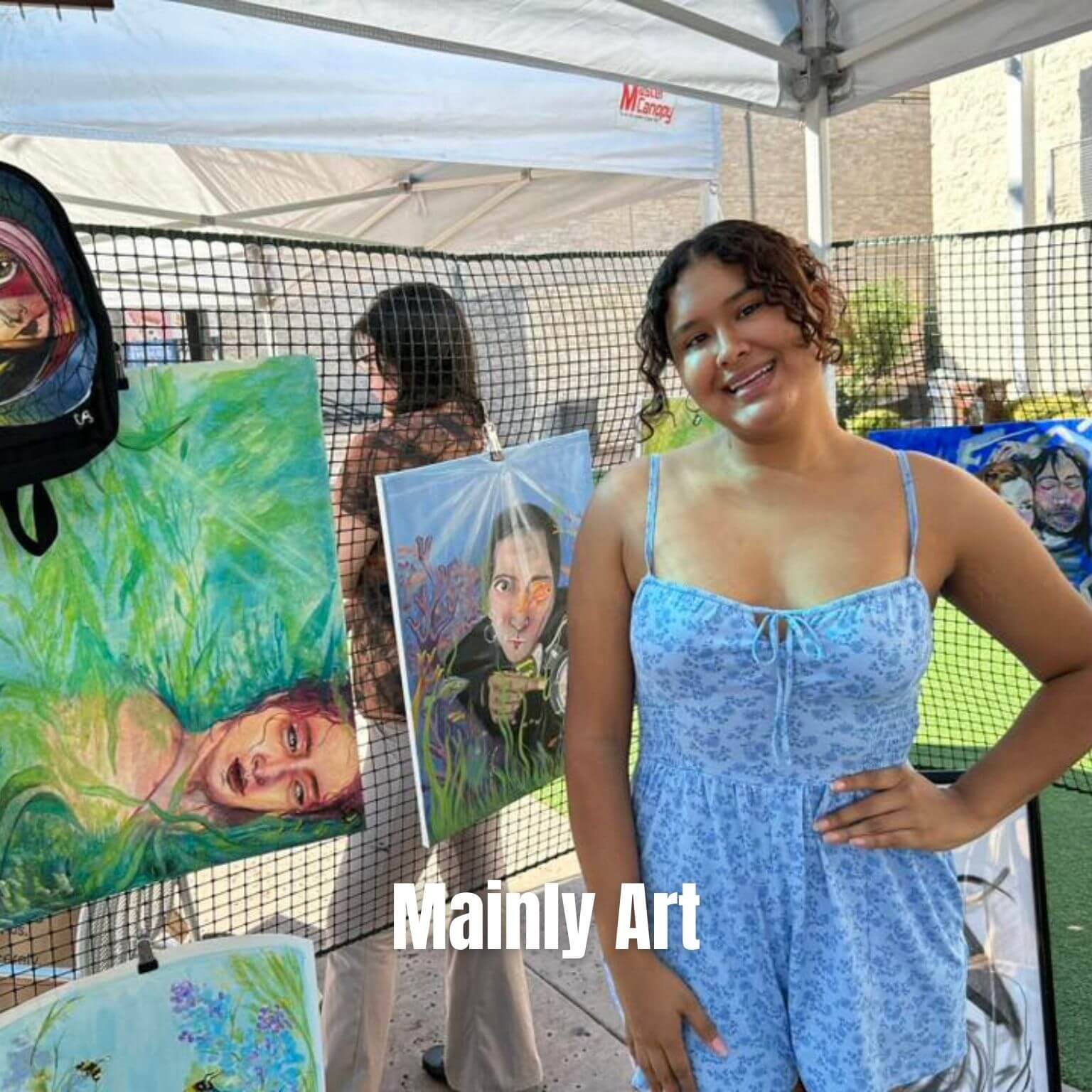 A young woman in a blue dress smiles while standing in front of colorful paintings at an outdoor art exhibit. Another person is visible in the background, and the words “Mainly Art” appear at the bottom.