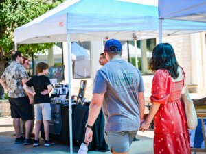 A couple holding hands walks toward a vendor booth at an outdoor market, while another group of people talk to the vendor under a white canopy.