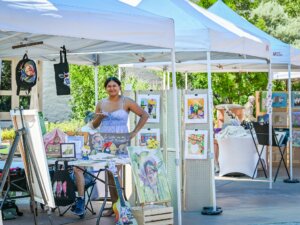 A woman stands at an outdoor art booth under a white canopy, surrounded by colorful paintings, framed artwork, and a painted guitar, on a sunny day.