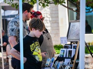 A child and two adults browse art prints and postcards at an outdoor market stall on a sunny day.