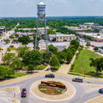 Aerial view of a roundabout with a Downtown sign, surrounded by greenery, parked cars, and buildings. A tall water tower labeled Round Rock stands prominently in the center background.