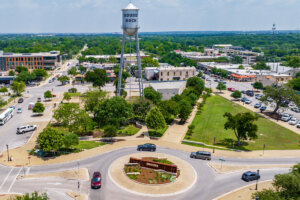 Aerial view of a roundabout with a Downtown sign, surrounded by greenery, parked cars, and buildings. A tall water tower labeled Round Rock stands prominently in the center background.