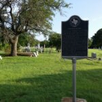 A historic marker stands in a grassy cemetery with several old tombstones and a large tree nearby, under a clear sky. The cemetery extends into the background with more headstones and trees.