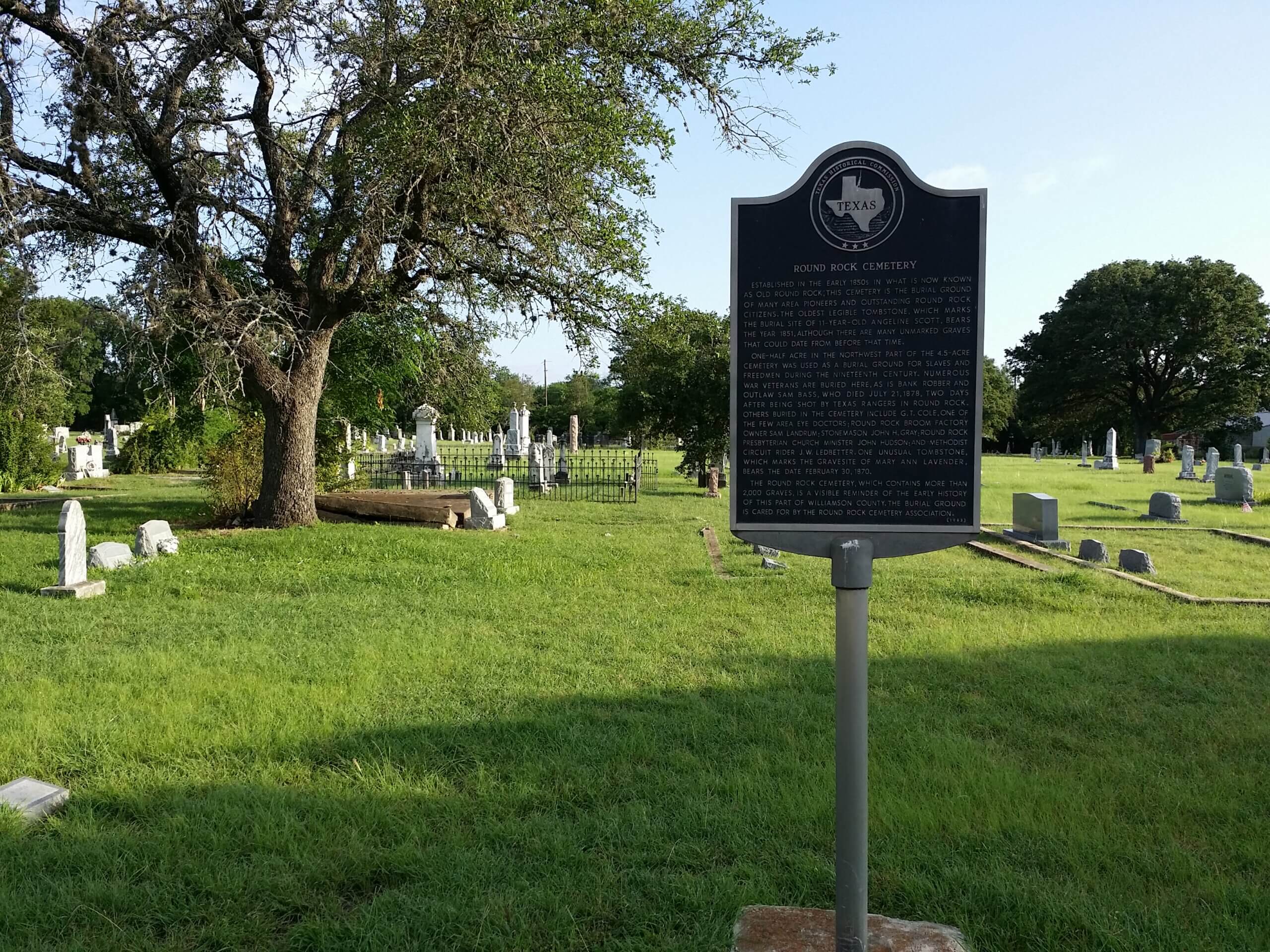 A historic marker stands in a grassy cemetery with several old tombstones and a large tree nearby, under a clear sky. The cemetery extends into the background with more headstones and trees.