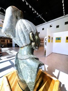 A stone sculpture of a woman’s head and shoulders with detailed hair and necklace is displayed on a wooden pedestal in a well-lit art gallery with white walls and various artworks in the background.