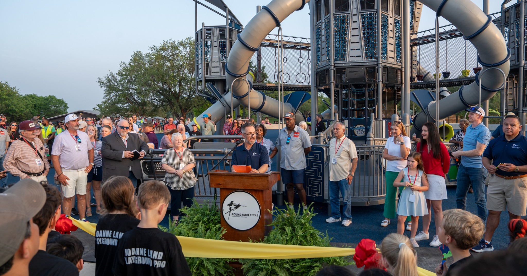 A large group gathers at an outdoor playground for a ribbon-cutting ceremony. A woman speaks at a podium while others stand nearby, with children and adults watching. The modern playground structure is visible in the background.