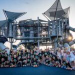 A large group of children and a few adults pose together in front of a modern playground structure with slides and towers on a sunny day. The children are wearing matching black shirts.