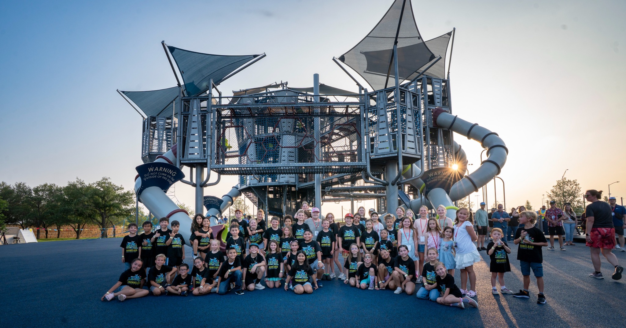 A large group of children and a few adults pose together in front of a modern playground structure with slides and towers on a sunny day. The children are wearing matching black shirts.