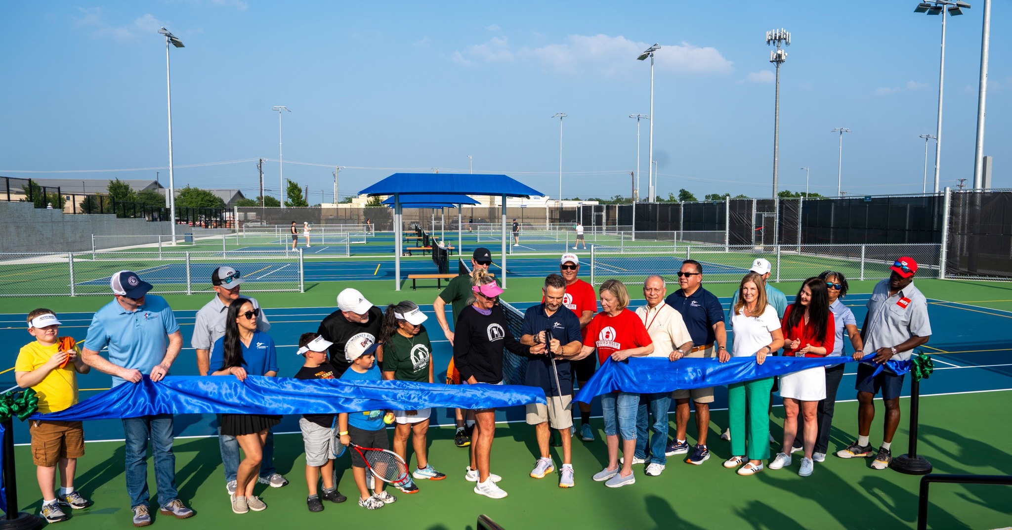 A group of people, including children and adults, stand in a line on a tennis court holding a long blue ribbon at a ribbon-cutting ceremony, with more tennis courts and clear sky in the background.