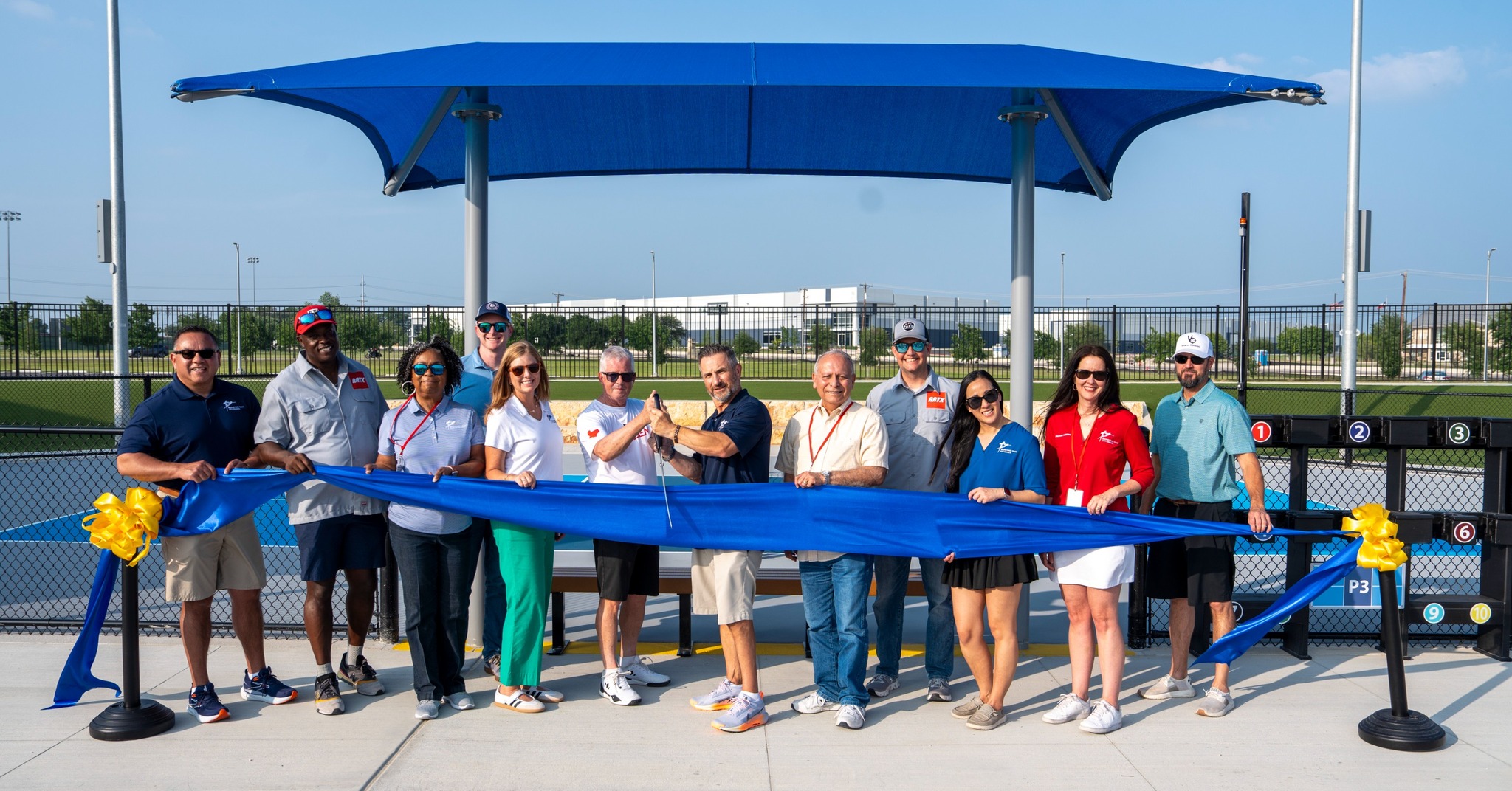 A group of people stand in front of a blue canopy, smiling and holding a large blue ribbon with bows at both ends, suggesting a ribbon-cutting ceremony at a sports facility.