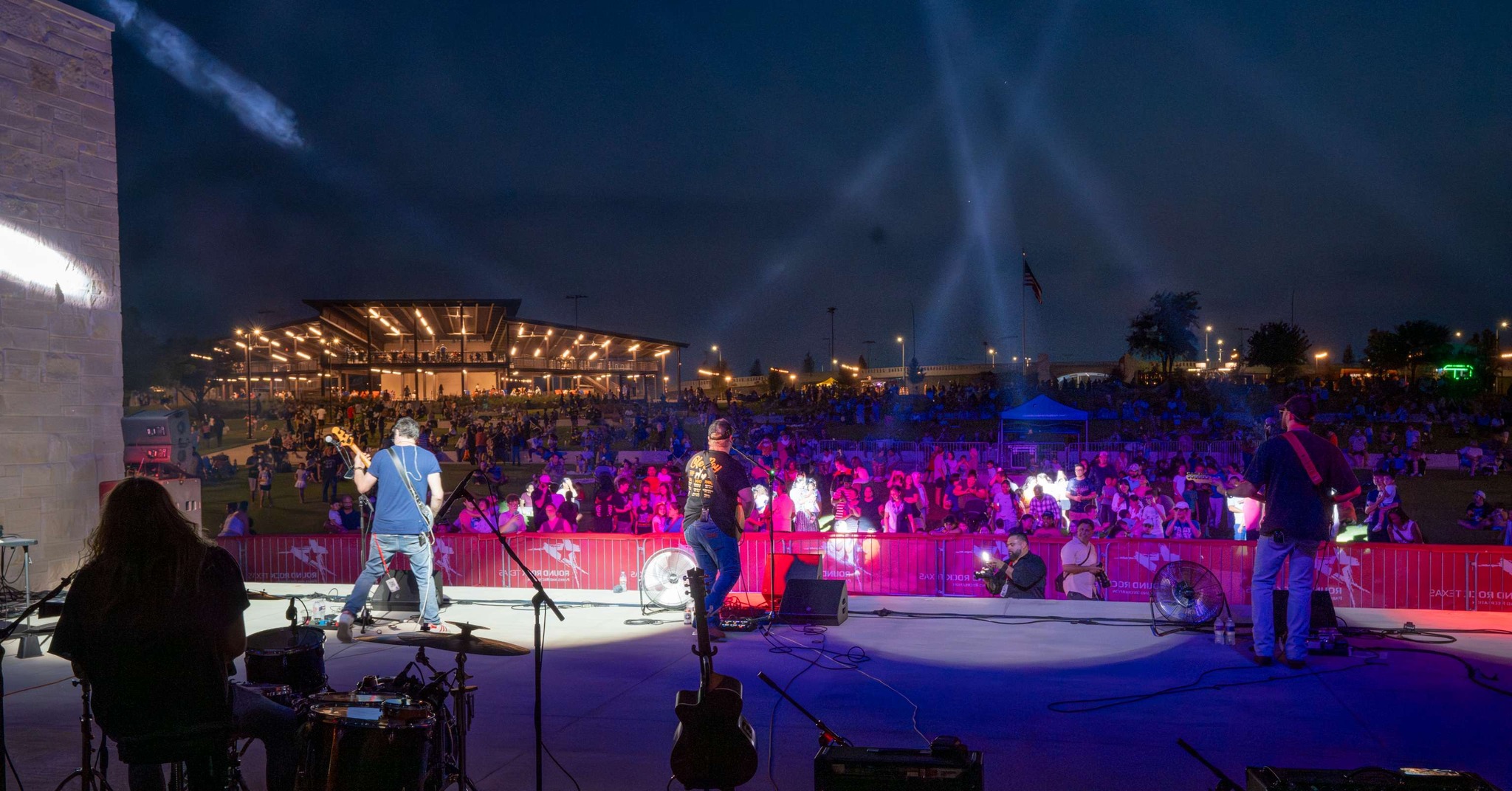 A band performs on an outdoor stage at night, facing a large, colorful crowd. Bright stage lights and beams illuminate the scene, with a pavilion and trees visible in the background.
