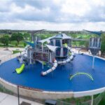 A modern playground with blue flooring features large climbing structures, multiple slides, tube tunnels, and shade canopies. The playground is surrounded by green trees and park facilities under a partly cloudy sky.