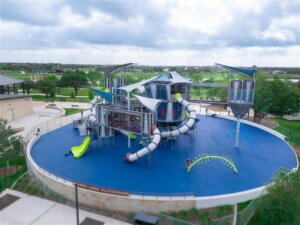 A modern playground with blue flooring features large climbing structures, multiple slides, tube tunnels, and shade canopies. The playground is surrounded by green trees and park facilities under a partly cloudy sky.