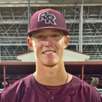 A young baseball player in a maroon uniform and matching cap with RR on the front stands smiling in front of a dugout with bleachers and teammates in the background.