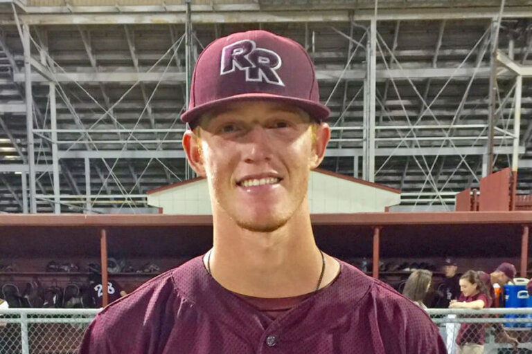 A young baseball player in a maroon uniform and matching cap with RR on the front stands smiling in front of a dugout with bleachers and teammates in the background.