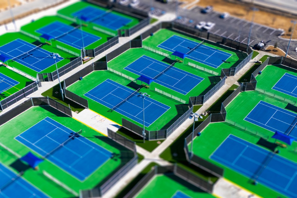 Aerial view of multiple blue and green tennis courts arranged in rows, separated by tall black fences, with nearby parking lots and pathways visible.