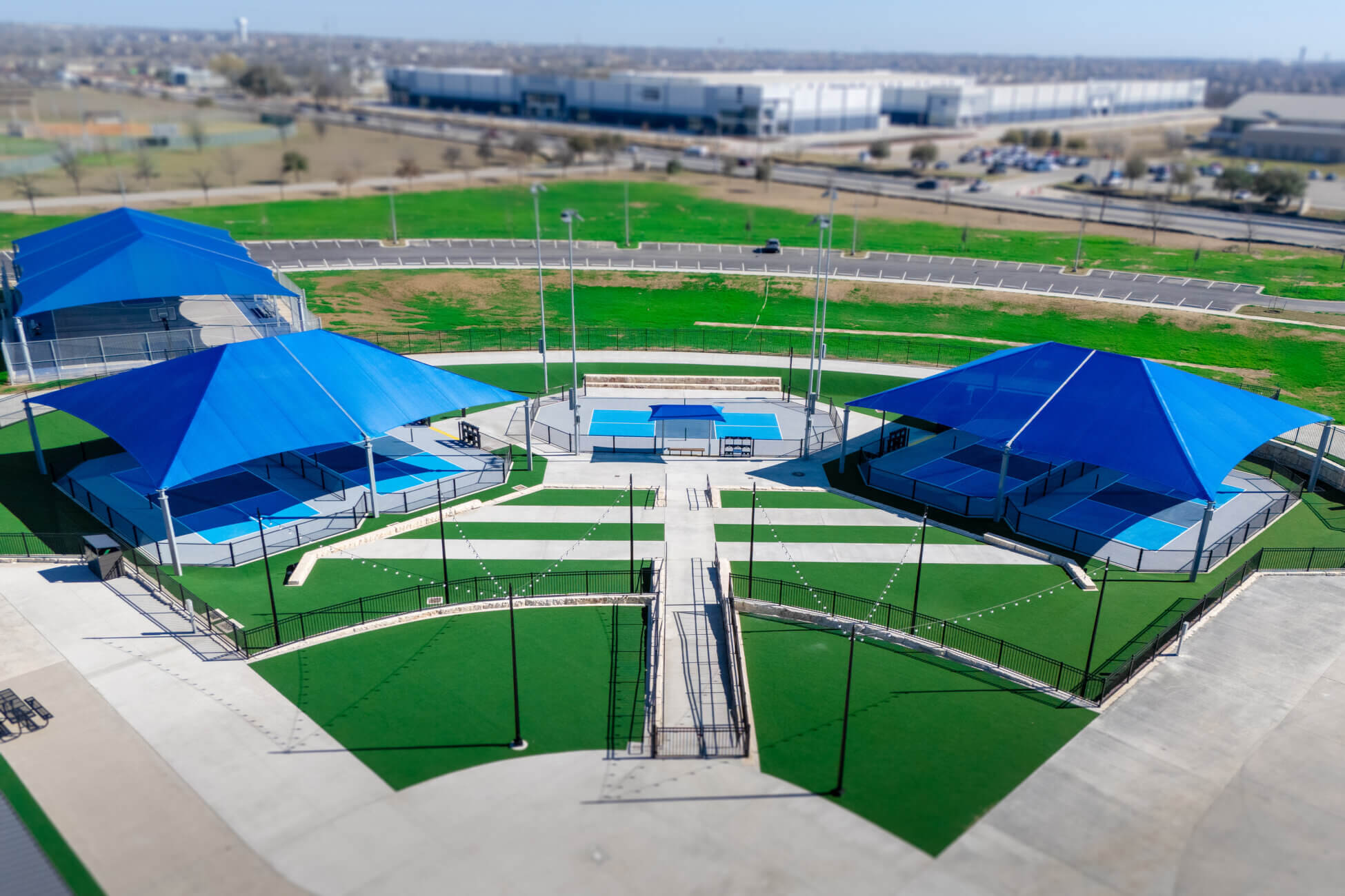 Aerial view of a sports complex with blue canopies covering several courts, surrounded by green artificial turf, fences, and concrete walkways, with buildings and open fields in the background.