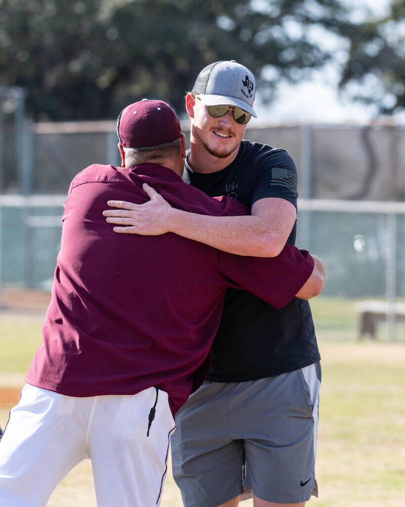 Two men hug on a sunny outdoor sports field; one wears a maroon cap and uniform, the other wears sunglasses, a gray cap, a black shirt, and gray shorts. Trees and a fence are visible in the background.