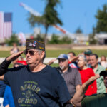 A group of people, including veterans in military hats, stand outdoors and salute. An American flag is visible in the background on a sunny day.