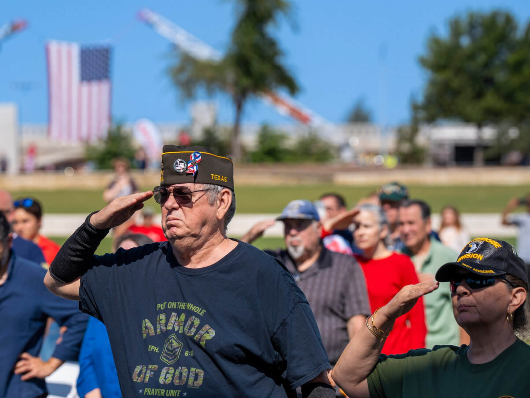 A group of people, including veterans in military hats, stand outdoors and salute. An American flag is visible in the background on a sunny day.
