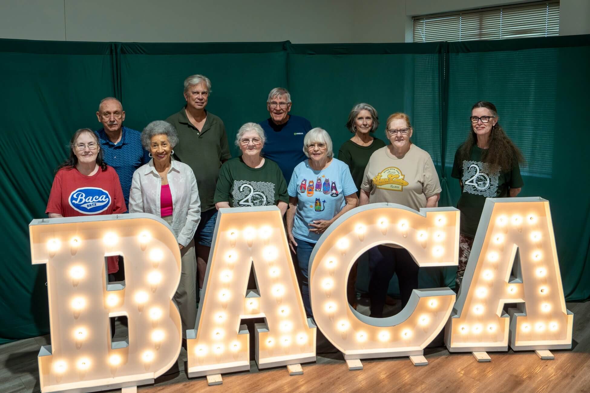 Ten smiling adults stand indoors behind large illuminated letters spelling BACA, with a green curtain and window in the background. Some wear t-shirts with logos and numbers, and all look at the camera.