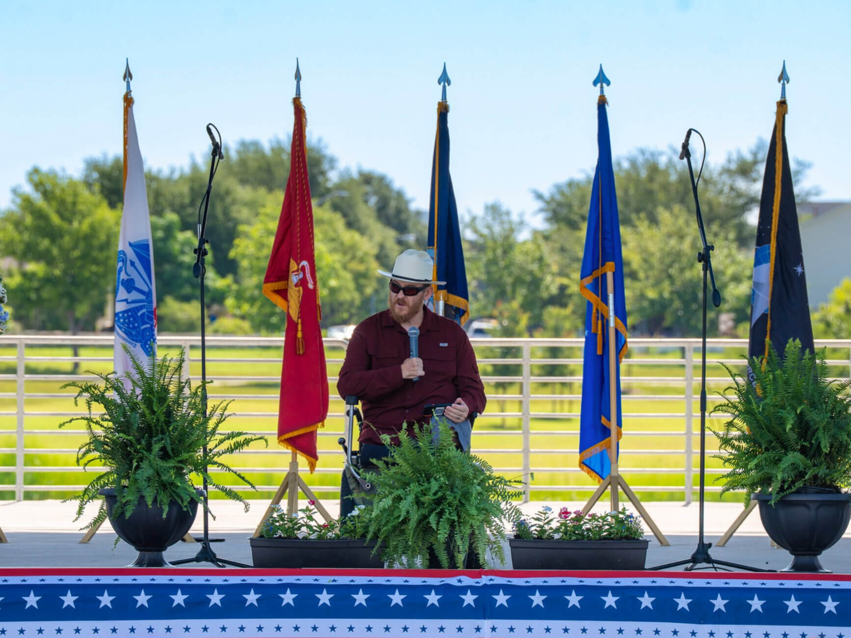 A man in a wheelchair speaks into a microphone on an outdoor stage decorated with potted ferns, American-themed bunting, and several military flags. Trees and grass are visible in the background.