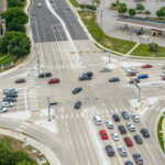 Aerial view of a busy multi-lane intersection with cars waiting at traffic lights and moving through the crossing; buildings and a parking lot are visible nearby.