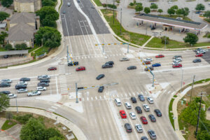 Aerial view of a busy multi-lane intersection with cars waiting at traffic lights and moving through the crossing; buildings and a parking lot are visible nearby.