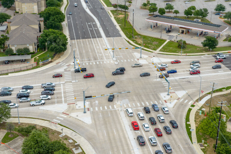 Aerial view of a busy multi-lane intersection with cars waiting at traffic lights and moving through the crossing; buildings and a parking lot are visible nearby.
