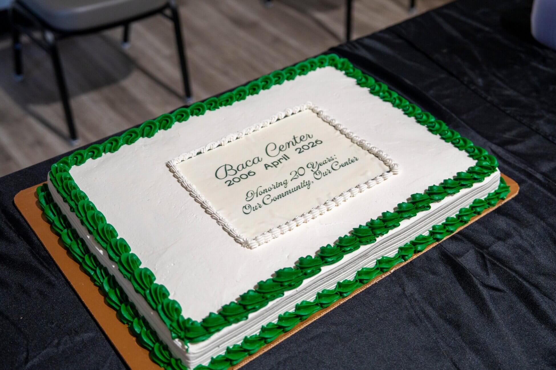A large rectangular cake with white icing and green borders sits on a table. The cake reads: Baca Center 2003-April 2023 Honoring 20 Years of Community and Events.