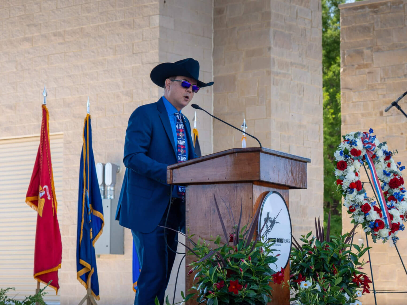 A man in a blue suit, cowboy hat, and sunglasses speaks at a podium decorated with plants and a patriotic wreath. Flags are displayed behind him against a stone wall background.