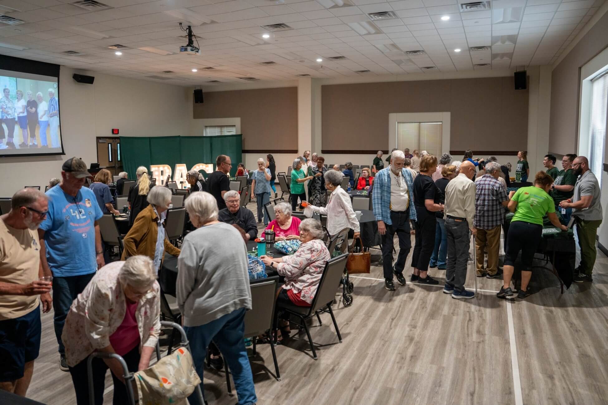 A large group of people, mostly older adults, gather in a community center room with tables and chairs. Some are seated and talking, while others stand and mingle. A slideshow of photos is projected on a screen in the background.