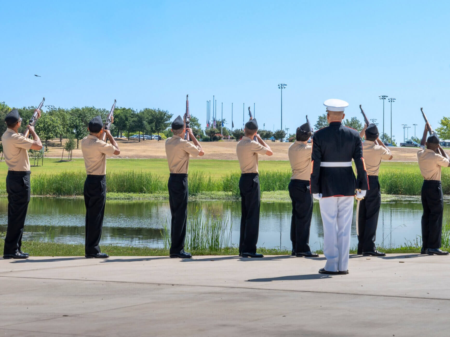 Seven uniformed individuals stand in a line by a pond, aiming rifles upward, with an officer in white observing. The scene appears to be a ceremonial or military honor guard outdoors.
