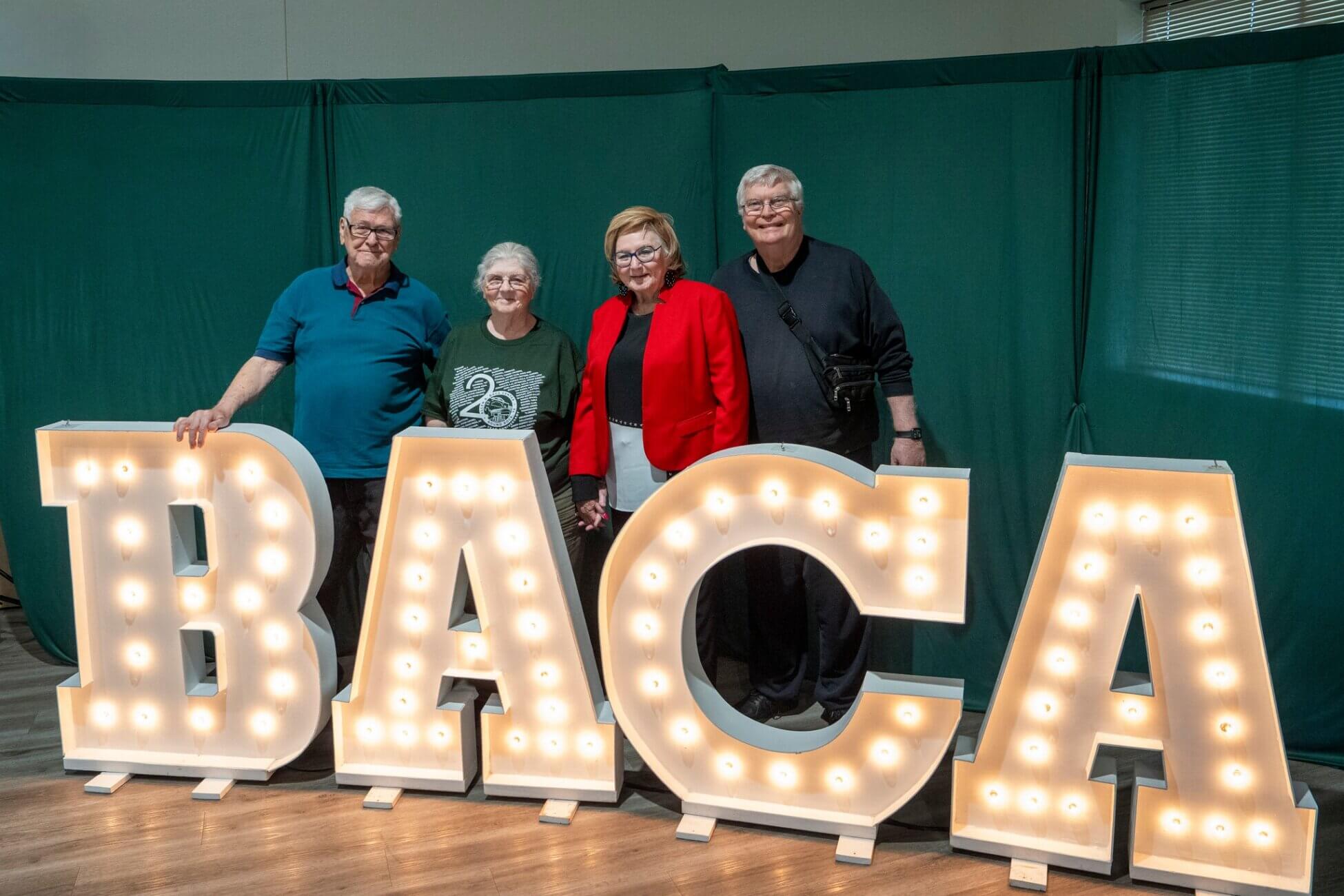 Four older adults stand smiling behind large, illuminated marquee letters spelling BACA with a green curtain backdrop.
