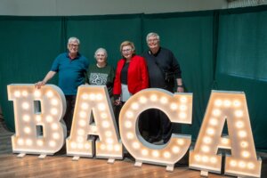 Four older adults stand smiling behind large, illuminated marquee letters spelling BACA with a green curtain backdrop.