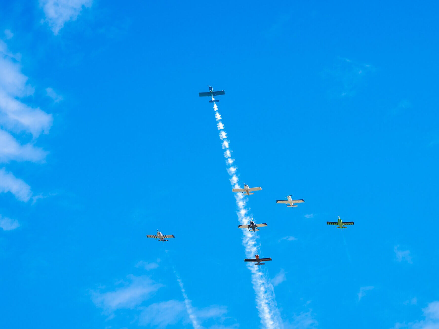 Six small airplanes fly in formation against a blue sky with wispy clouds. One plane in the center leaves a trail of white smoke as it ascends higher than the others.