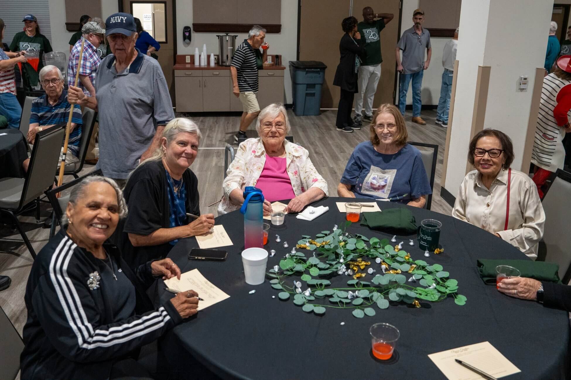 A group of six older adults sit around a round, decorated table, smiling at the camera during a social gathering. Other people are visible in the background, mingling in a large room.