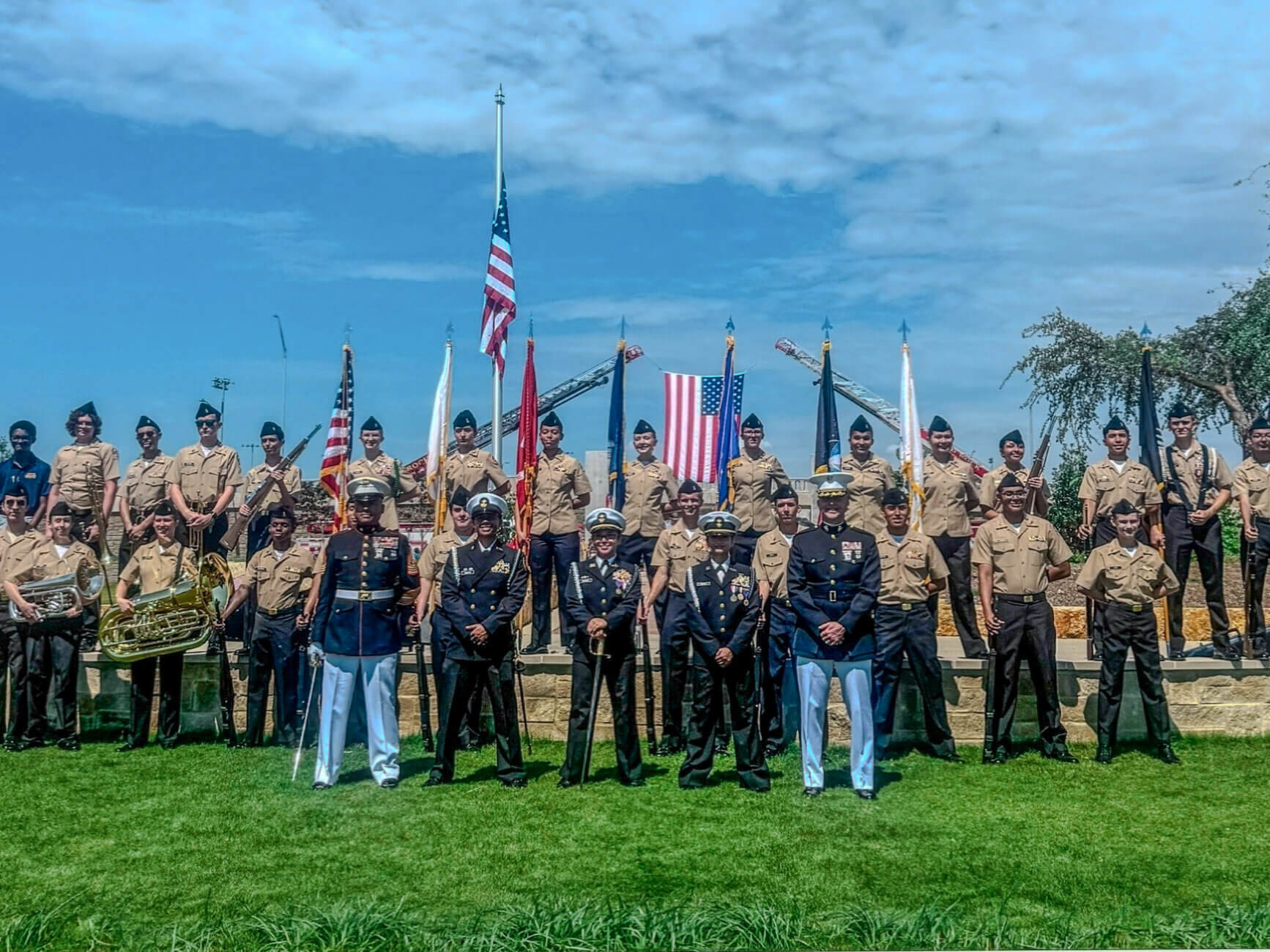 A group of uniformed military cadets poses on a grassy field, holding various flags, including the American flag. Some members play band instruments. A large American flag is displayed in the background under a partly cloudy sky.